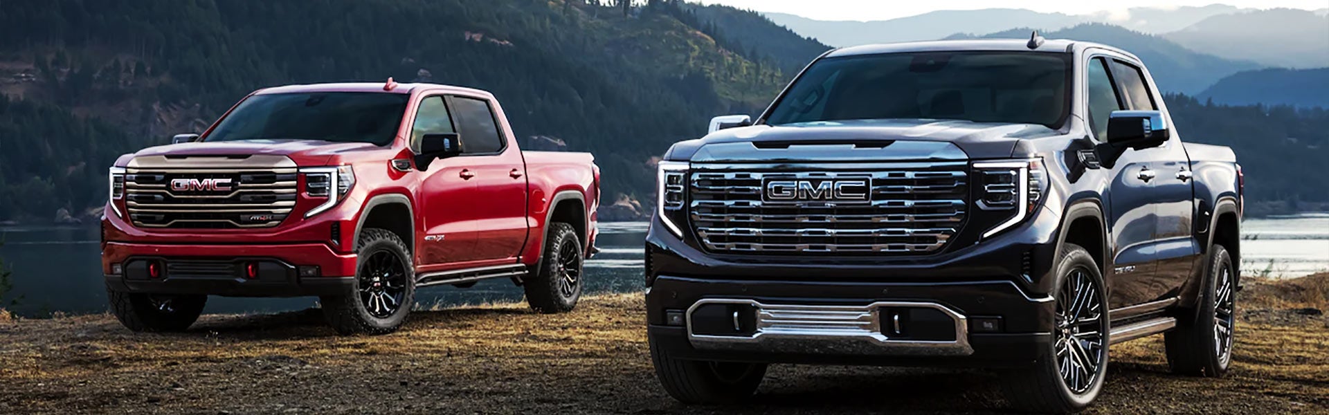 Two GMC Sierra 1500 trucks parked side by side with a mountain and pond in the background